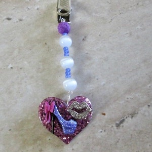Puede incluir: Un charm en forma de corazón rosa con un zapato de tacón alto morado brillante, un lazo rosa y un diseño de labios morados brillantes. El charm está unido a un clip plateado y una cadena de cuentas blancas y moradas.