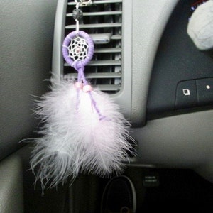 May include: A purple and white dreamcatcher hanging from the rearview mirror of a car. The dreamcatcher has white feathers and is attached to a silver chain.