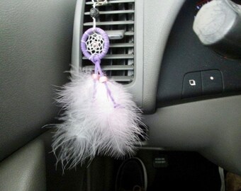 May include: A purple and white dreamcatcher hanging from the rearview mirror of a car. The dreamcatcher has white feathers and is attached to a silver chain.