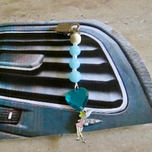 Puede incluir: Un ambientador de coche azul y plateado con un amuleto en forma de corazón y una figura de hada. El amuleto está hecho de vidrio facetado y el hada está hecho de metal.