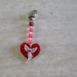 Puede incluir: Un amuleto de corazón rojo con un diseño de ángel plateado cuelga de un clip plateado con cuentas rojas y transparentes.