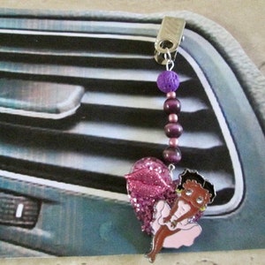 Puede incluir: Un ambientador de coche rosa y morado con un personaje de dibujos animados de una mujer con cabello negro y un vestido rosa. El ambientador está sujeto a una rejilla de ventilación del coche con un clip plateado y una cadena de cuentas.