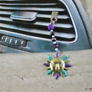 Puede incluir: Un clip para la ventilación del coche con un colgante de sol dorado, detalles en azul y morado, y una cadena de cuentas. La cadena tiene cuentas moradas y marrones. El clip es plateado.