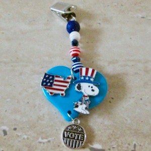 May include: A blue heart-shaped charm with a red, white, and blue American flag and a cartoon dog wearing a red, white, and blue hat. The charm is attached to a clip with red, white, and blue beads and a silver charm that says "VOTE".