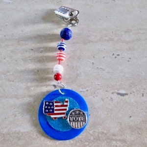 May include: A blue and silver charm with a red, white, and blue beaded chain. The charm features a small American flag and the word "VOTE" in a banner.