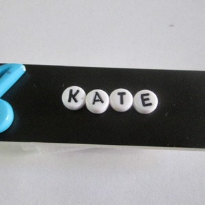 May include: A black name tag with a blue musical note and the name "KATE" spelled out in white letters.