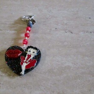 May include: A black heart-shaped charm with a red glitter finish features a cartoon character in a red dress and a red lip. The charm is attached to a silver clip with red beads.