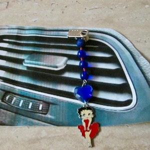 Puede incluir: Un amuleto para la ventilación del coche de color azul y rojo con una mujer de dibujos animados con un vestido rojo y un corazón azul. El amuleto está unido a una cuerda de cuentas azules.