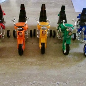 Puede incluir: Una fila de cinco motos de juguete en rojo, naranja, amarillo, verde y azul. Cada moto tiene un asiento negro y un manillar plateado. Las motos están alineadas sobre una superficie gris.