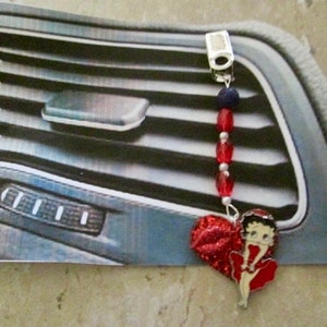 Puede incluir: Un amuleto de coche rojo y blanco con un corazón rojo y un personaje de dibujos animados con un vestido rojo. El amuleto está sujeto a una rejilla de ventilación de coche.