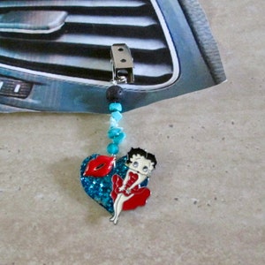 Puede incluir: Un amuleto de ambientador de coche azul y rojo con un personaje de dibujos animados y un diseño en forma de corazón. El amuleto está unido a un clip plateado y una cadena de cuentas.