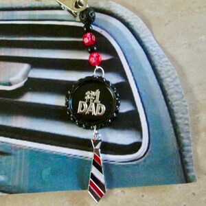 Puede incluir: Un ambientador de coche negro y plateado con una cadena de cuentas rojas y negras. El dije presenta las palabras "#1 DAD" en letras plateadas. El dije está unido a una corbata a rayas blancas y negras.