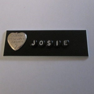 May include: A black rectangular sign with the name "JOSIE" spelled out in white letters on black tiles. A silver heart charm with the inscription "Sorry I'm ready to make a difference" is on the left side of the sign.