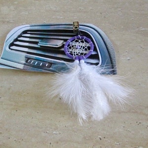 May include: A purple and white dreamcatcher with white feathers hanging from it. The dreamcatcher is attached to a car's air vent.