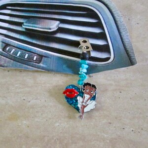Puede incluir: Un ambientador de coche azul y rojo con un personaje de dibujos animados y un diseño de labios rojos. El ambientador está colgado de una rejilla de ventilación del coche.