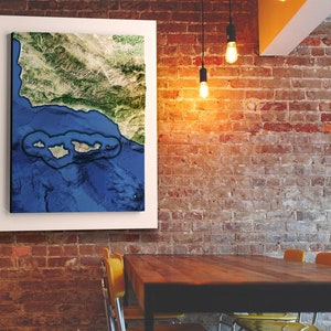 May include: A framed canvas print of a satellite image of a coastline with islands. The image is in shades of blue, green, and brown. The canvas is hanging on a brick wall in a room with a wooden table and chairs.