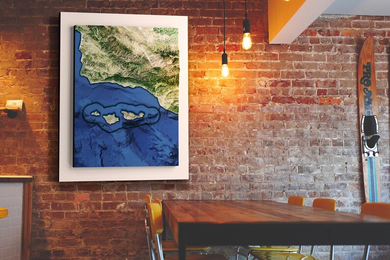 May include: A framed canvas print of a satellite image of a coastline with islands. The image is in shades of blue, green, and brown. The canvas is hanging on a brick wall in a room with a wooden table and chairs.