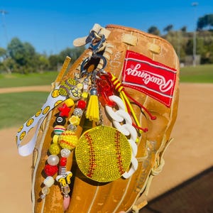 May include: A tan Rawlings baseball glove with a decorative keychain. The keychain features a yellow rhinestone softball, beaded charms, a white chain, and a yellow tassel. The background is a baseball field.