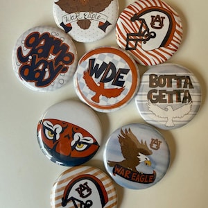 Puede incluir: Una colección de ocho chapas redondas con temas de la Universidad de Auburn. Los diseños incluyen águilas, cascos de fútbol americano y textos como War Eagle, Game Day, WDE y Botta Getta en paletas de colores naranja, azul, blanco y marrón.