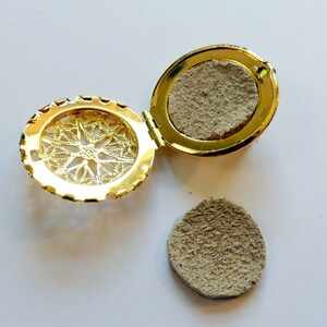 May include: A gold locket with a filigree design on the front and a round, beige felt pad inside. The locket is open and the felt pad is sitting next to it.