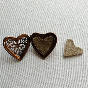 May include: Open copper-colored heart-shaped locket with filigree design, revealing a brown interior. A small, separate heart-shaped piece of tan material is also present. The locket is on a white surface.