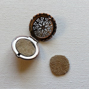 May include: A round, open locket with a filigree design. The locket is made of metal and has a brown finish. The locket is open and the inside is lined with a brown felt material. There is a separate piece of brown felt material next to the locket.