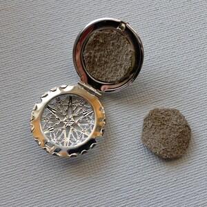 May include: A silver locket with a filigree design and a brown felt pad inside. The locket is open and the pad is visible. There is a separate brown felt pad next to the locket.