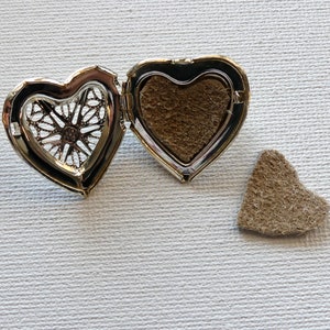 May include: Open silver heart-shaped locket with a brown felt insert. The locket is open and the felt insert is visible. There is a separate brown felt heart-shaped piece next to the locket.