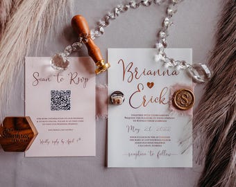 Puede incluir: Un conjunto de invitaciones de boda con una tarjeta blanca con texto en cobre que dice Brianna y Erick, la fecha 21 de mayo de 2022 y un sello de cera con la letra S. Una tarjeta RSVP separada incluye un código QR, un bloque hexagonal de madera.