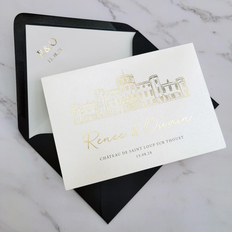 May include: A cream-colored wedding invitation card featuring a gold foil illustration of a chateau, the names Renee & Owain, and the text CH&Acirc;TEAU DE SAINT LOUP SUR THOUET 15.08.28. The card is tucked into a black envelope with R & O 15.08.28 in gold.