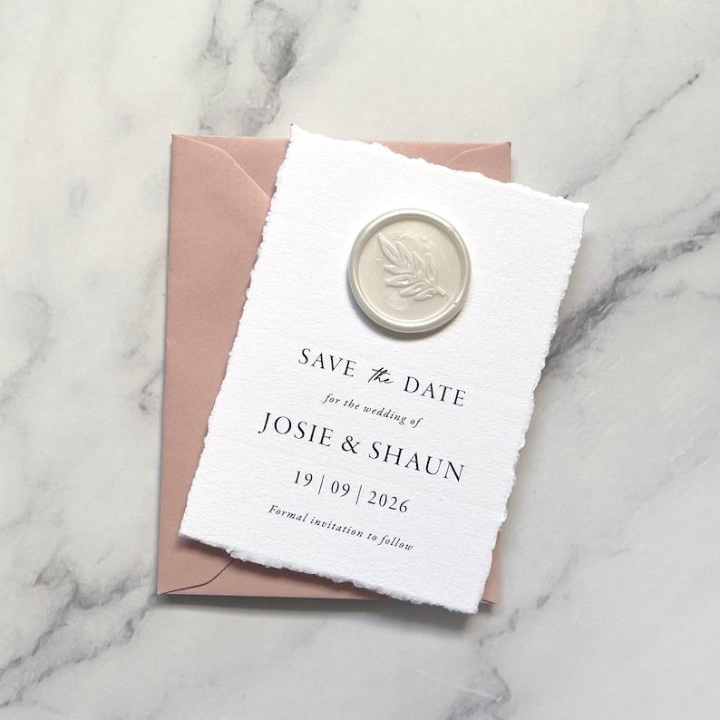 May include: A white save the date card with a white wax seal with a leaf design. The card has black text that reads "SAVE the DATE for the wedding of JOSIE & SHAUN 19 | 09 | 2026 Formal invitation to follow."
