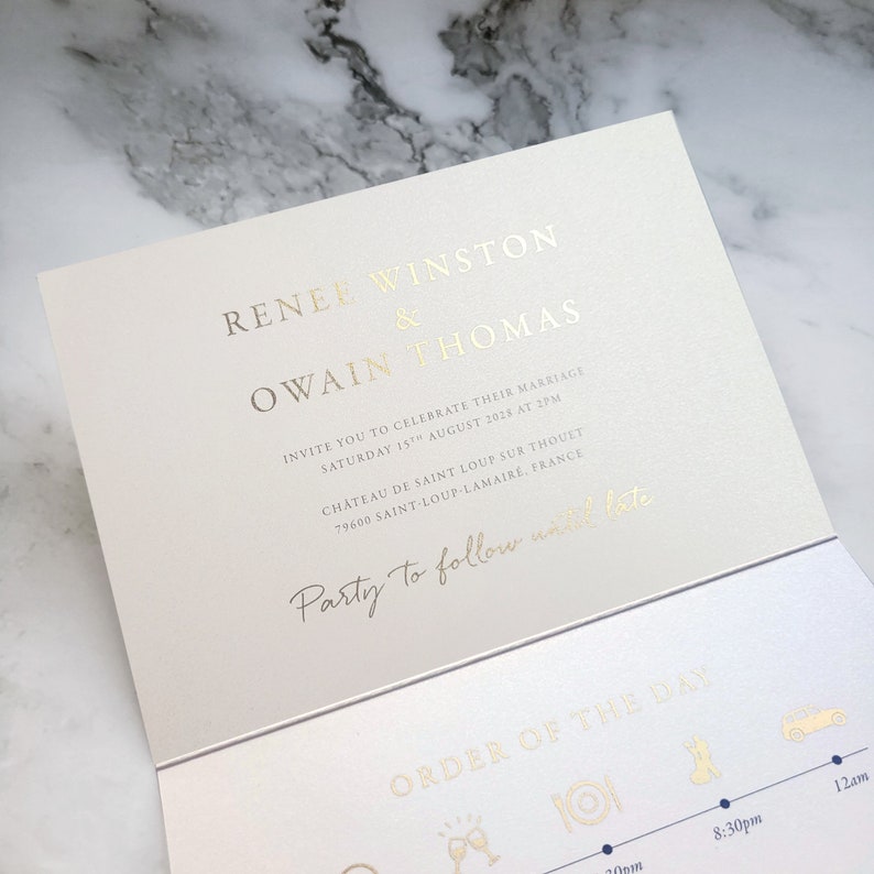 May include: A cream-colored wedding invitation card featuring gold foil lettering. The text reads Renee Winston & Owain Thomas, invite you to celebrate their marriage, Saturday 15th August 2028 at 2pm, Ch&acirc;teau de Saint Loup sur Thouet, Party to follow until late, and Order of the Day.
