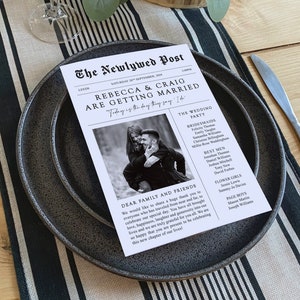 以下が含まれることがあります： 「The Newlywed Post」と題された、新聞のようにデザインされた結婚式のメニュー。白黒のデザインには、カップルの写真、名前、結婚式のパーティーの詳細、感謝のメッセージが掲載されています。メニューは濃い灰色のプレートに置かれています。