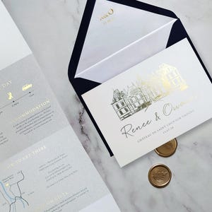 May include: A white wedding invitation card with gold foil architectural illustration and the text Renee & Owain, Chateau de Saint Loup sur Thouet, 15.07.28. Includes a dark blue envelope with gold R&O text and two gold wax seals with leaf imprints.