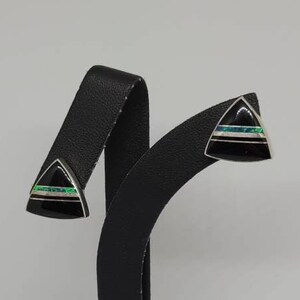 May include: A pair of black triangle-shaped earrings with green and white opal inlay. The earrings are displayed on a black stand.
