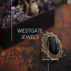 K&ouml;nnte beinhalten: Ein goldener Ring mit einem gro&szlig;en schwarzen Edelstein, der von kleineren, klaren Edelsteinen umgeben ist. Der Ring liegt auf einer Holzoberfl&auml;che mit dem Text "WESTGATE JEWELS" und "VINTAGE JEWELRY".