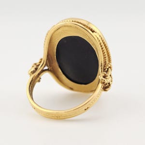 May include: A gold ring featuring an oval, black gemstone. The ring has an intricate, rope-style design around the stone and on the shoulders of the band. The background is plain and light.