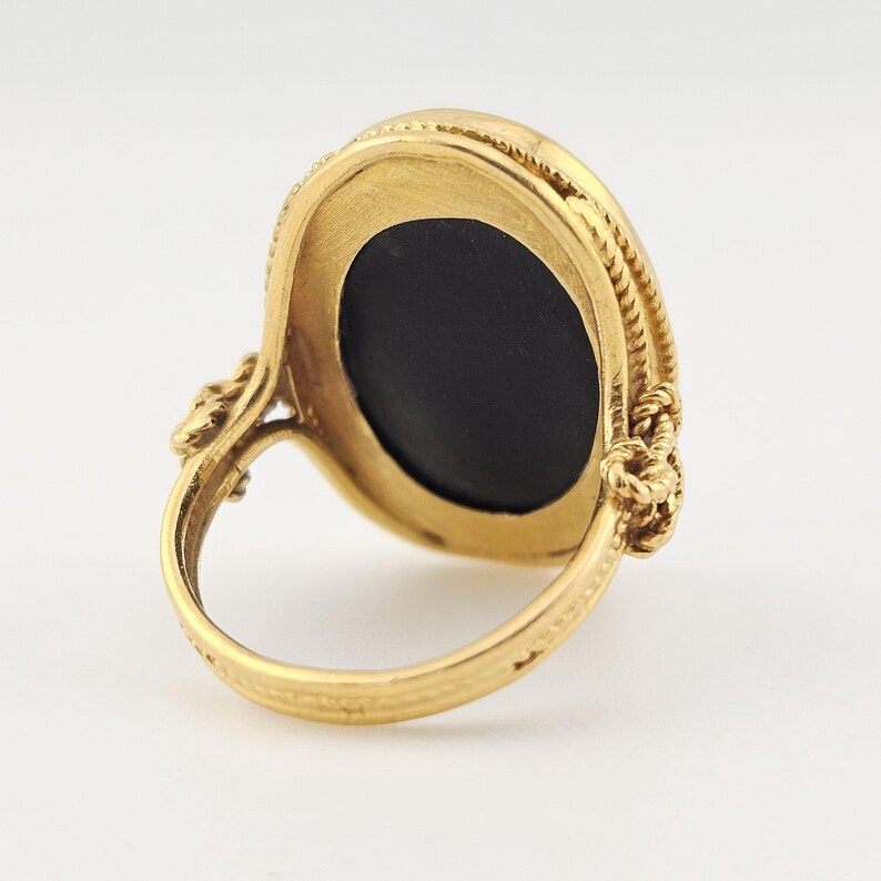 May include: A gold ring featuring an oval, black gemstone. The ring has an intricate, rope-style design around the stone and on the shoulders of the band. The background is plain and light.