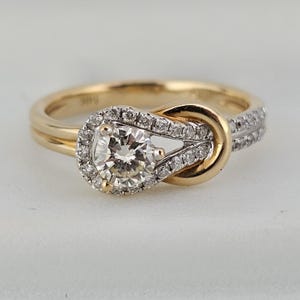 May include: A gold engagement ring with a knot design. The ring features a large, round, clear diamond at the center, surrounded by a halo of smaller diamonds. Additional small diamonds are set along the band.