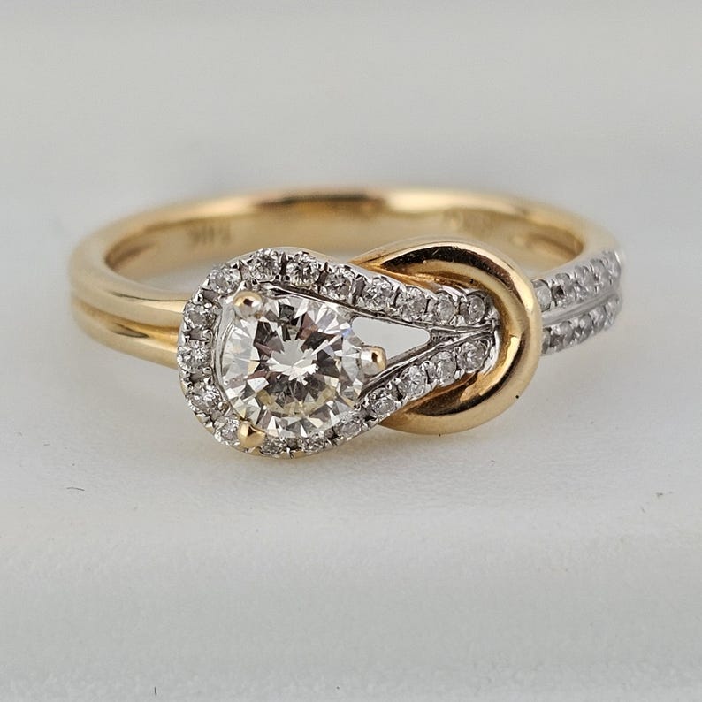 May include: A gold engagement ring with a knot design. The ring features a large, round, clear diamond at the center, surrounded by a halo of smaller diamonds. Additional small diamonds are set along the band.