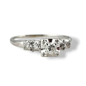 May include: A vintage silver ring with a square-cut center diamond and smaller accent stones on each side. The ring has a delicate band and a classic, timeless design, perfect for an engagement or special occasion.