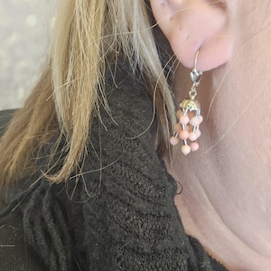 May include: A dangling earring with a silver-colored metal top and a cluster of small, round, peach-colored beads. The earring is attached to the ear with a lever-back clasp. The earring is worn by a person with blonde hair.