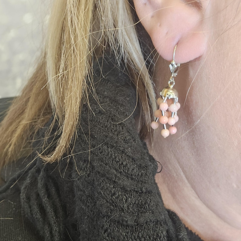 May include: A dangling earring with a silver-colored metal top and a cluster of small, round, peach-colored beads. The earring is attached to the ear with a lever-back clasp. The earring is worn by a person with blonde hair.