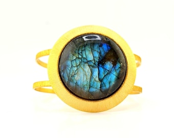 May include: A gold-toned ring featuring a large, circular, cabochon-cut labradorite gemstone. The stone displays iridescent blue and dark grey tones with visible internal fractures. The band consists of two thin, parallel gold-toned metal wires.