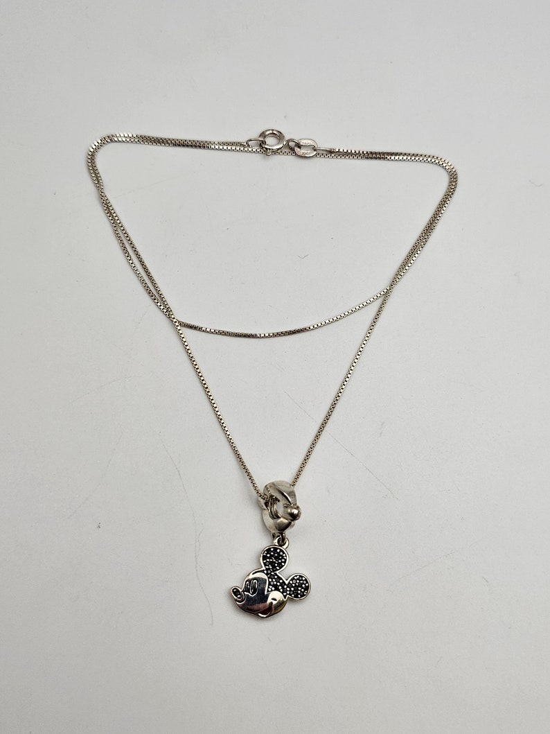 May include: A silver necklace featuring a Mickey Mouse pendant. The pendant has a black stone design for the ears and a silver face. The necklace chain is a delicate box chain style.