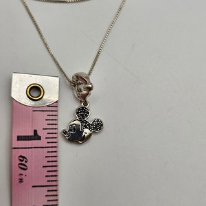 May include: Silver necklace featuring a Mickey Mouse charm with black accents. The charm is approximately 1 inch in height. The necklace is shown with a pink and white measuring tape.