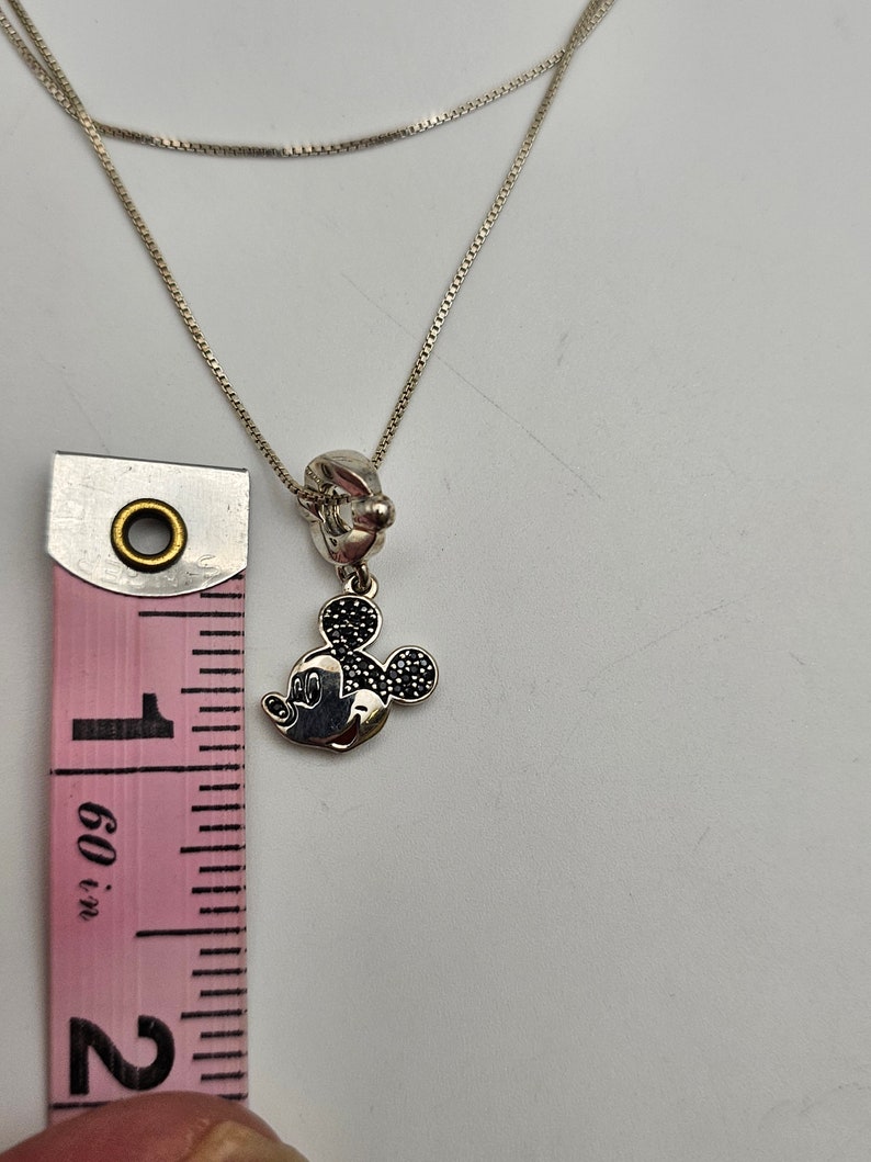 May include: Silver necklace featuring a Mickey Mouse charm with black accents. The charm is approximately 1 inch in height. The necklace is shown with a pink and white measuring tape.