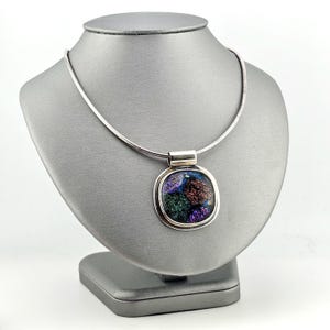Puede incluir: Un collar de tono plateado con un colgante rectangular de bordes redondeados y una incrustación de vidrio iridiscente oscuro. El vidrio muestra destellos brillantes en colores verde, morado y cobre. El colgante cuelga de una cadena plateada.