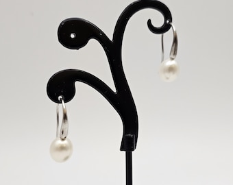 May include: A pair of silver hoop earrings with white pearl drops hanging from them. The earrings are displayed on a black earring stand.