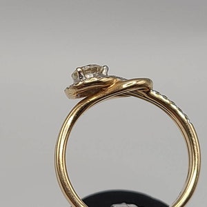 May include: A gold ring with a diamond center stone and smaller diamonds set around it. The ring has a twisted band design. The diamonds sparkle against the gold setting. The ring is shown against a neutral background.